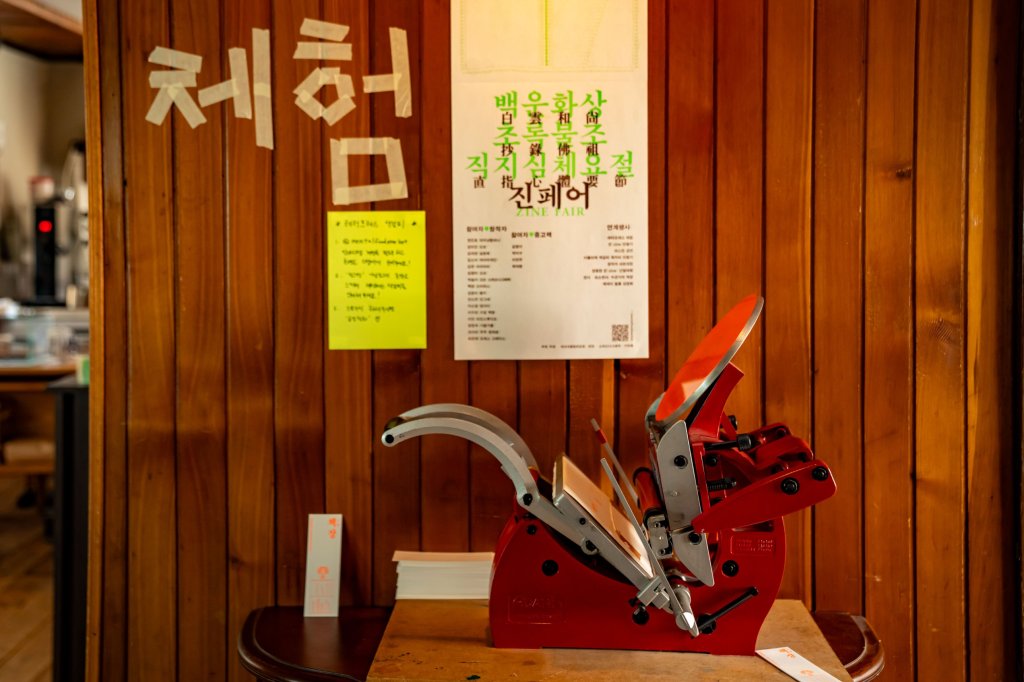 레터프레스 체험 — 테이프로 쓴 ‘체험’ 글자와 진페어 포스터 아래 빨간 레터프레스 인쇄기, 옆에 찍어낸 책;장 책갈피