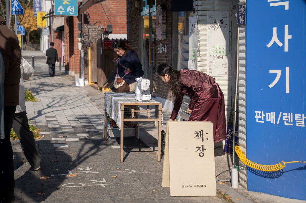 거리에서 재봉틀을 올린 테이블을 준비하는 사람들과 책;장 입간판