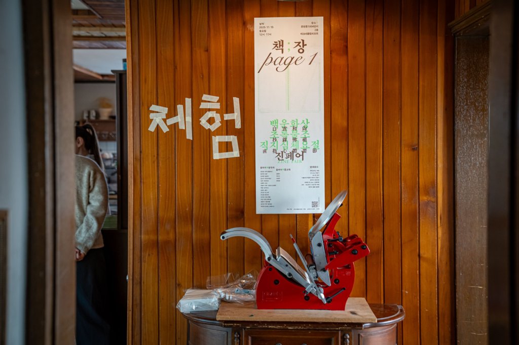 나무 벽에 테이프로 쓴 ‘체험’ 글자와 포스터 아래 놓인 빨간 레터프레스 인쇄기