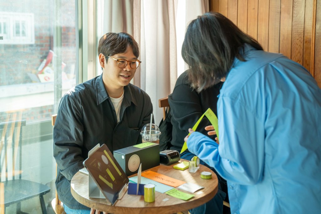 라임색 자와 문구를 놓은 동그란 테이블에서 방문객과 이야기하는 창작자