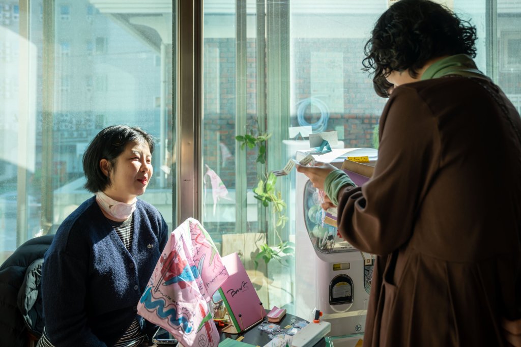 햇살 드는 창가 부스에서 방문객과 이야기하는 창작자