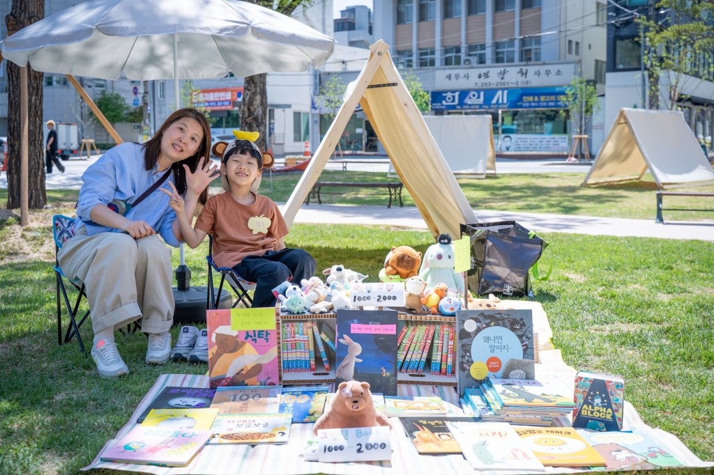 텐트 앞에서 인형과 그림책을 1,000–2,000원에 파는 어린이 셀러와 엄마