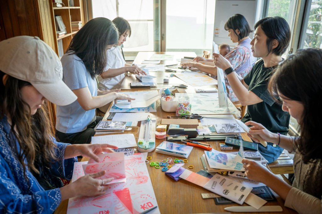리소 인쇄물과 제본 도구가 펼쳐진 긴 테이블에서 각자의 진을 접는 참여자들