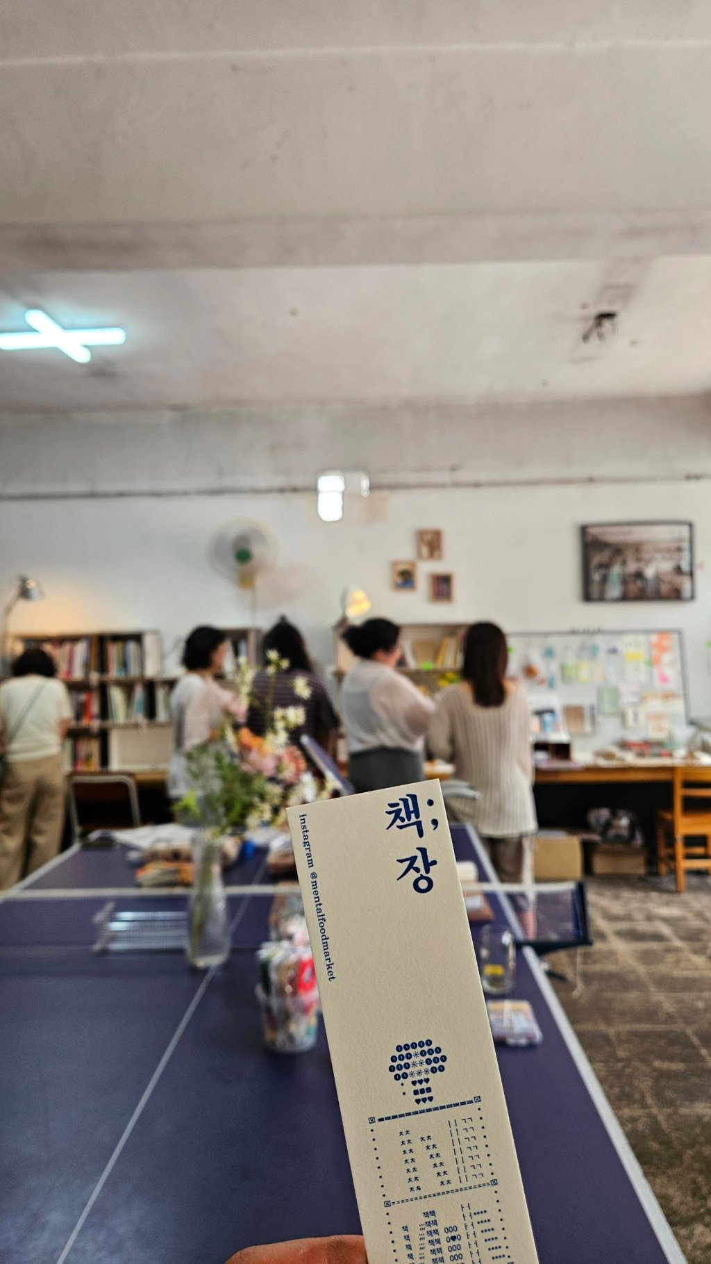 책;장 책갈피를 들고 있는 손 너머로 책장을 둘러보는 방문객들