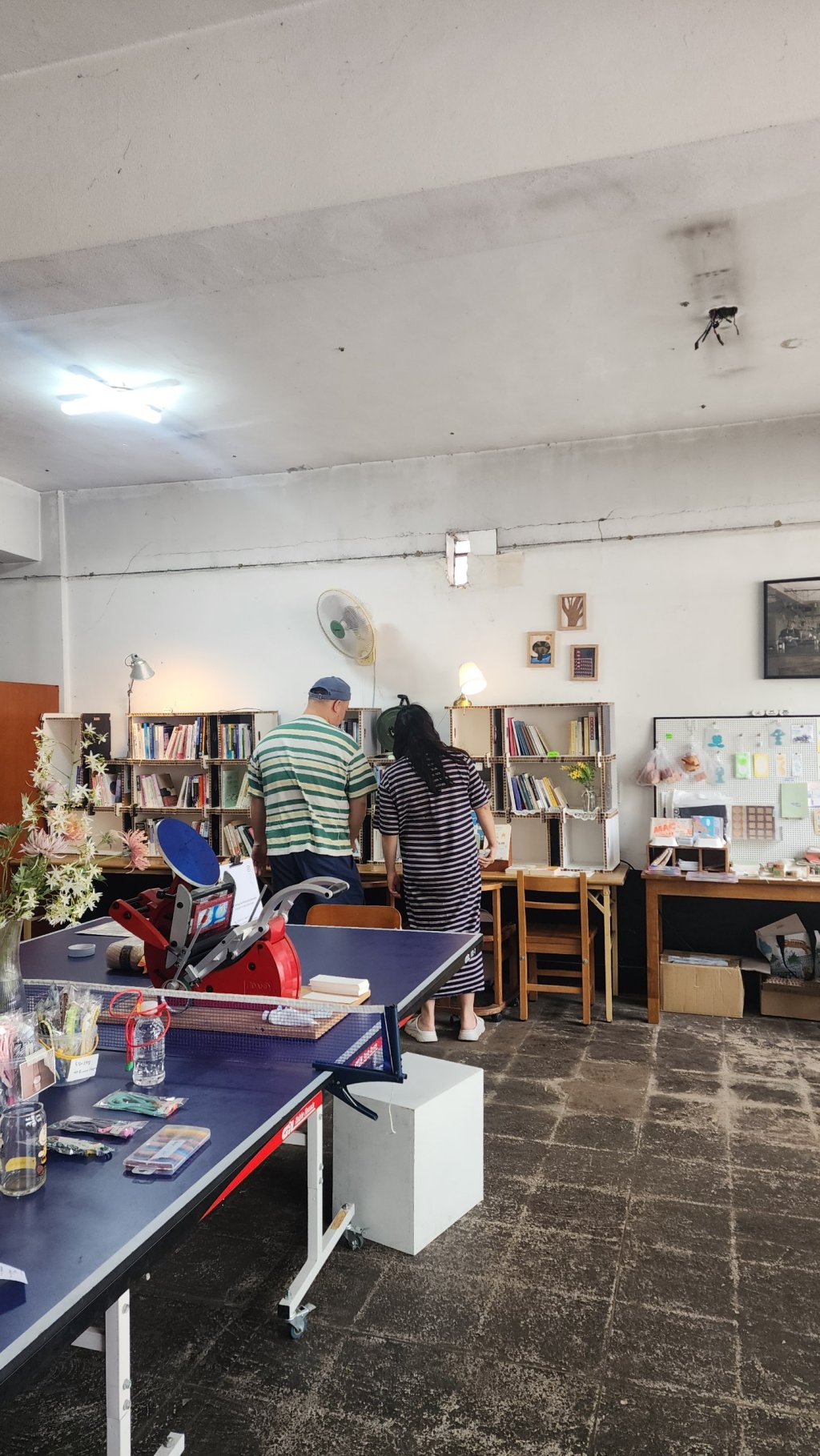 탁구대 위 레터프레스 인쇄기 뒤로 두 방문객이 책장을 살펴보는 모습